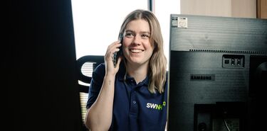 Eine junge Frau mit dem Logo SWNH sitzt lächelnd an einem Schreibtisch und telefoniert.