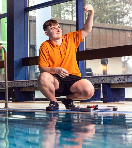 Ein junger Mann mit orangenem T-Shirt kniet neben einem Schwimmbecken und betrachtet eine Wasserprobe.