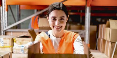 Eine junge Frau mit orangener Warnweste arbeitet lächend in einem Lager mit Kartons und Boxen.