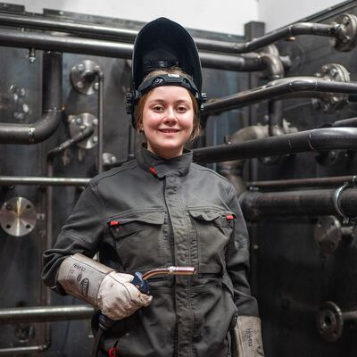 Eine junge Frau steht lächelnd in Arbeitskleidung neben einer komplexen Anlage mit verschiedenen Rohrleitungen.