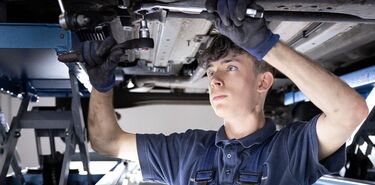 Ein junger Mann arbeitet mit Werkzeug an der UNterseite eines Kraftfahrzeugs.