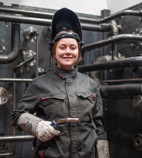 Eine junge Frau steht lächelnd in Arbeitskleidung neben einer komplexen Anlage mit verschiedenen Rohrleitungen.