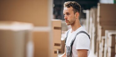 Ein junger Mann in Arbeitskleidung steht in einer Halle mit vielen Paketen und Boxen.