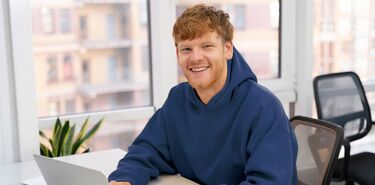 Ein junger Mann in einem blauen Pullover sitzt lächeln an einem Laptop.