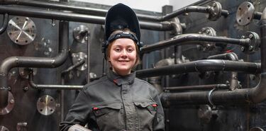 Eine junge Frau steht lächelnd in Arbeitskleidung neben einer komplexen Anlage mit verschiedenen Rohrleitungen.
