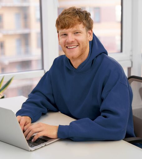 Ein junger Mann in einem blauen Pullover sitzt lächeln an einem Laptop.