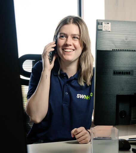 Eine junge Frau mit dem Logo SWNH sitzt lächelnd an einem Schreibtisch und telefoniert.